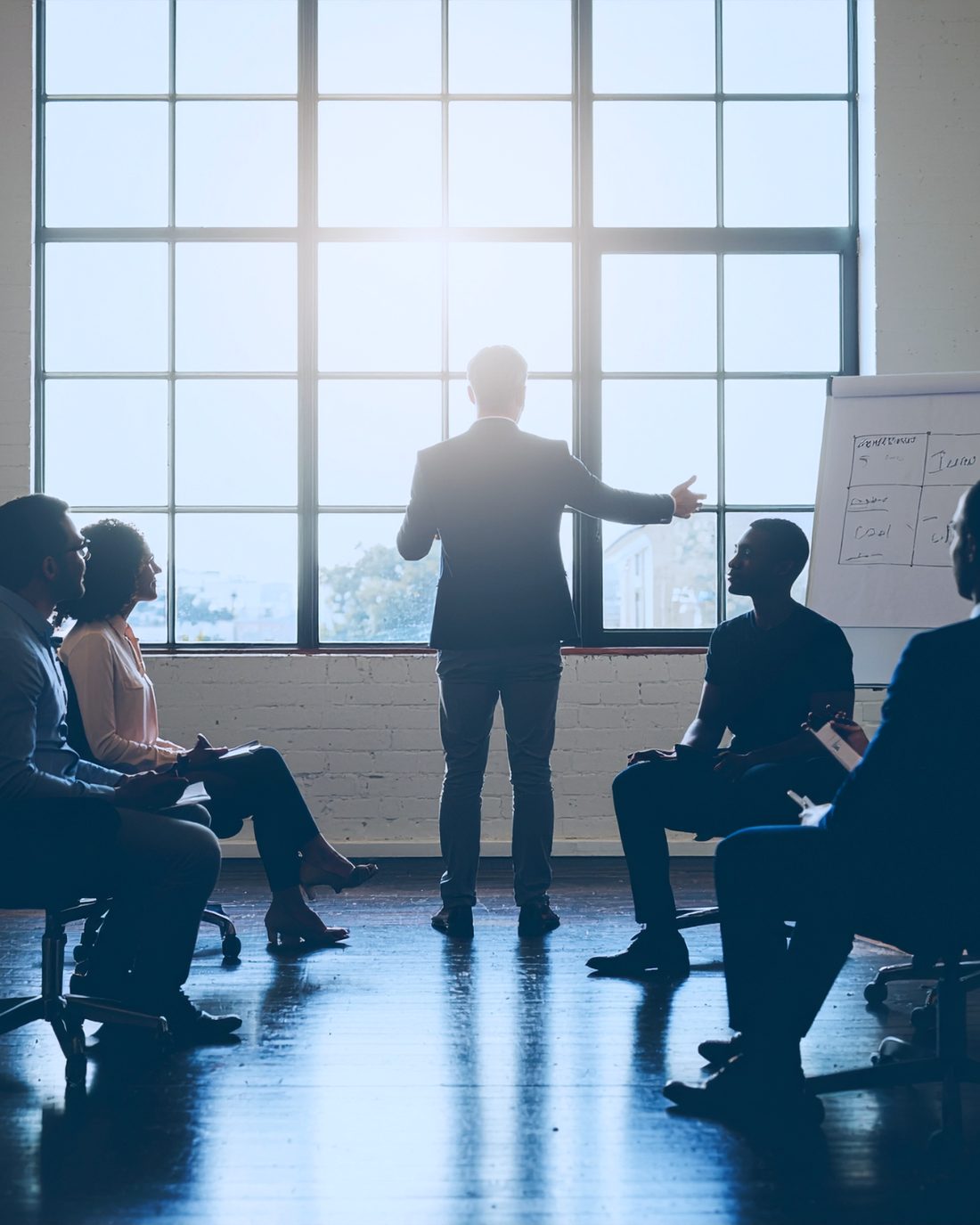 A facilitator standing at a large window leads a small group workshop, gesturing toward a flipchart.